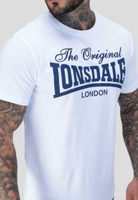 Mees, kellel on tätoveeritud käed ja kael, kannab valget T-särki, mille ees on tumesinised kiri "The Original Lonsdale London".