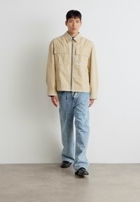 Jachetă bej cu fermoar în față, având două buzunare pe piept, asortată cu pantaloni din denim bleu deschis. Pantofi negri completează ținuta. Design simplu și utilitar.