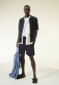 Navy gestructureerd kortemouwen shirt over een witte overhemd met knopen en navy shorts, terwijl een lichtblauw shirt wordt vastgehouden. Zwarte slidesandalen maken de outfit compleet.