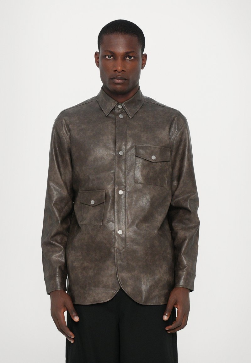Chemise en simili cuir marron avec un col pointu, fermeture à boutons sur le devant et deux poches poitrine. Présente une texture lisse et un ourlet arrondi.