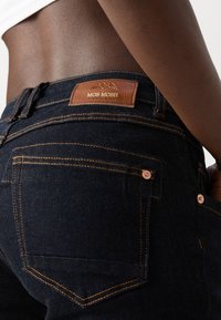 Jean en denim bleu foncé avec couture orange porté par une personne à la peau foncée, montrant la poche arrière et l'étiquette en cuir marron MOS MOSH.