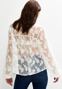 Witte doorschijnende blouse met bloemenborduursels, lange mouwen en een losse pasvorm. De achterkant toont een vloeiend ontwerp, gelaagde textuur en gedetailleerde stiksels.