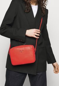 Rød læder skuldertaske med rektangulær form, lynlås og justerbar rem; præget logo foran. Båret med en sort blazer.