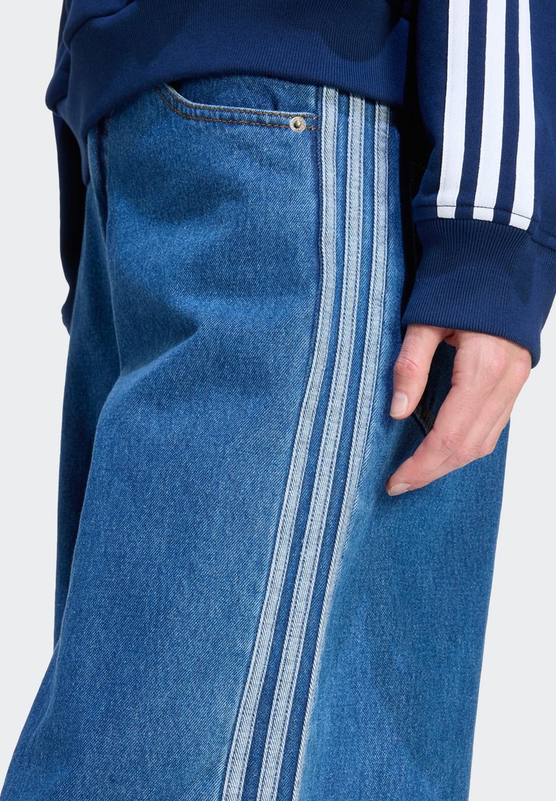 Pantalon en denim à jambes larges bleu moyen avec des bandes latérales blanches contrastantes, présentant une texture subtilement usée et une poche frontale.