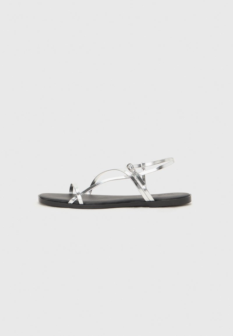 Sølvfargede metall sandaler med et minimalistisk design, som har tynne stropper og en sort flat såle. Fremhevingene inkluderer kryssremmer.