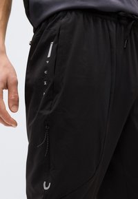 Κοντινή λήψη μαύρου αθλητικού παντελονιού Puma με πλαϊνή τσέπη με φερμουάρ και ελαστική μέση, φορεμένο από άτομο με ορατό αριστερό χέρι.