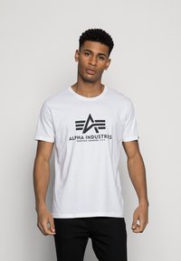 T-shirt alb din bumbac cu un logo grafic negru, cu litere stilizate și trei linii orizontale pe față. Mâneci scurte și guler rotund.