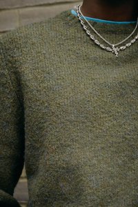 Olivgrön stickad tröja med en texturerad yta, med rund halsringning och lager av silverkedjor med intrikata detaljer.