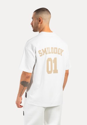 Hvid bomulds t-shirt med ryg grafik "SMILODOK 01" i beige. Korte ærmer, afslappet pasform og synlige syninger ved kanten.