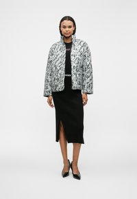 Mels un balts abstrakts raksts ar gaismu biezinātu virsjakas ar augstu apkakli un sānu kabatām, kas kombinēts ar melnu ribotu svārku un augstpapēžu kurpēm.