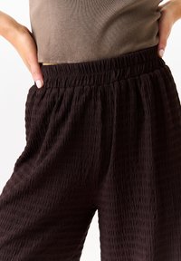Black textured pants with an elastic waistband; fabric has a crinkled appearance. Model partially visible, wearing a fitted brown top.