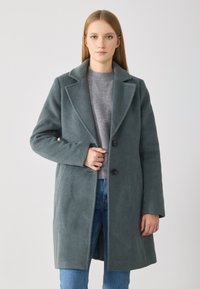 Cappotto grigio in lana con colletto a risvolto, chiusura con bottoni singoli e tasche laterali, indossato sopra un maglione grigio chiaro e jeans blu.