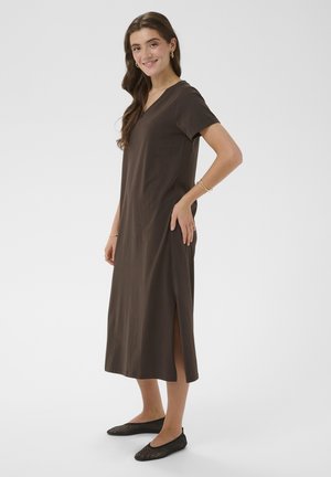 Uśmiechnięta kobieta w brązowej sukience midi z dekoltem w serek, rozcięciem po bokach i czarnych plecionych balerinach, stojąca bokiem na jednolitym tle.