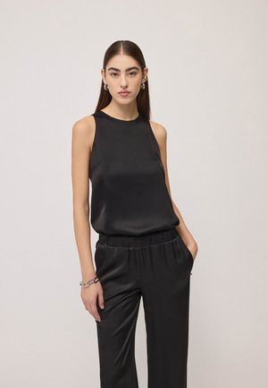 Vrouw met lang donker haar, gekleed in een zwarte satijnen mouwloze top en bijpassende zwarte satijnen broek met één hand in de zak.