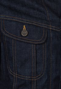 Mörk denimjacka med kontrasterande kopparfärgade sömmar, ficklock och en metall-LEE-logotyp. Texturerat tyg med en klassisk design.