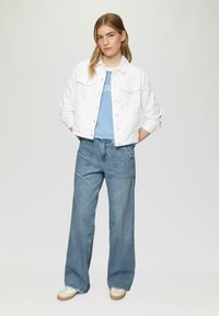 Veste en denim blanche, T-shirt rayé bleu, jeans légers bleus amples et chaussures blanches. Le mannequin se tient les mains dans les poches, sur un fond neutre.