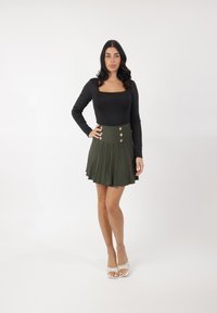 Body nero a maniche lunghe abbinato a una gonna plissettata verde scuro con vita alta e sei bottoni dorati come decorazione. Scarpe bianche con punta aperta.