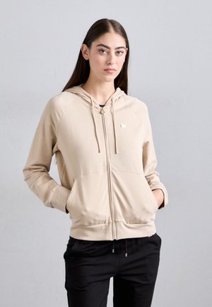 Beige rits-hoodie van zachte stof, met een capuchon, twee voorzakken en een gouden logo detail. Gecombineerd met zwarte broek.