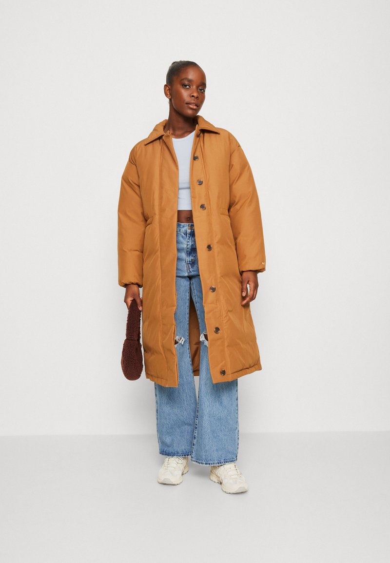 Braune Puffermantel mit Kragen, Knopfverschluss und seitlichen Taschen, kombiniert mit einem hellblauen Crop-Top und weitem Hosenstil. Strukturierter brauner Accessoire.