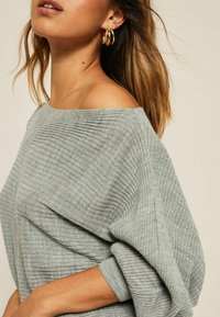 Maglione grigio a coste scollato con vestibilità ampia e maniche larghe. Presenta un design a maglia testurizzata e un neckline rilassato. Orecchini a cerchio d'oro.