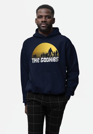 Mand iført navy hættetrøje med en gul sol og silhuetter af syv figurer over teksten "The Goonies".