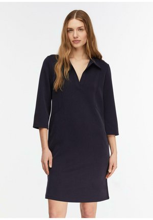 Kobieta z długimi włosami w ciemnogranatowej sukience do kolan z rękawami do łokcia i dekoltem w serek, stojąca na jednolitym tle.