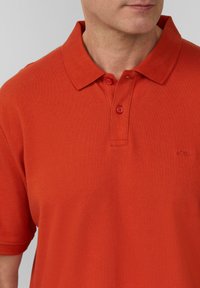 Oranges Polohemd aus strukturiertem Baumwollmaterial, mit klassischem Kragen, drei Knöpfen und einem gestickten Logo auf der Brust.