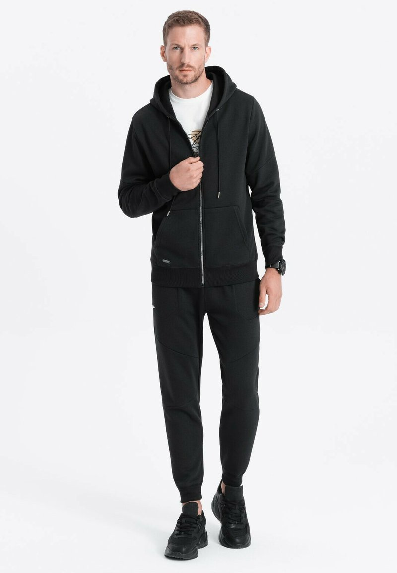 Sort lynlås hoodie med en justerbar hætte, parret med mørke, tapered joggers med sømdetaljer. Bæres med sorte sneakers.