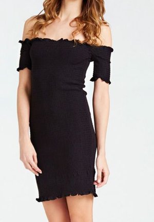 Kobieta ubrana w dopasowaną, czarną sukienkę odsłaniającą ramiona z krótkimi rękawami i falbanami na krawędziach, stojąca na jednolitym białym tle.