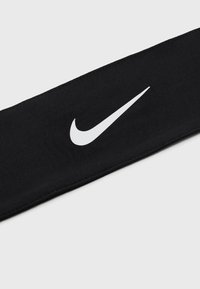 Crna tkanina traka za glavu s glatkom teksturom i elastičnim rastezanjem, sa istaknutim bijelim Nike Swoosh logotipom.