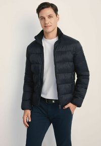 Czarna kurtka puchowa z poziomymi przeszyciami, wysokim kołnierzem i kieszeniami na zamek. Noszona na białej koszulce i w zestawie z ciemnymi spodniami.