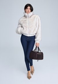 Giacca in finta pelliccia color crema chiaro con colletto alto, abbinata a jeans aderenti blu scuro, tacchi a punta color nude e una piccola borsa marrone.