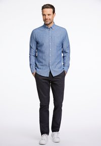 Camicia azzurra chiara con bottoni e tasca, abbinata a pantaloni scuri e sneaker bianche, su uno sfondo bianco semplice.