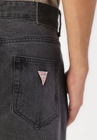 Melna denima bikses ar aizmugurējo kabatu, kurā ir trīsstūrveida sarkans un balts "GUESS" logotipa plāksne. Redzama tekstūru josta un šuvju detaļas.