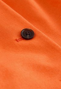 Gros plan d'un bouton marron foncé cousu sur un tissu orange vif avec une couture visible autour de la boutonnière.