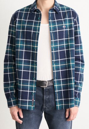 Person trägt ein aufgeknöpftes blau-grünes kariertes Hemd über einem weißen Tanktop, dunkle Jeans und einen schwarzen Gürtel mit silberner Schnalle.