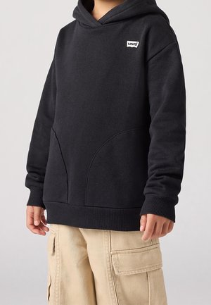 Bambino che indossa una felpa con cappuccio nera Levi's con tasche frontali e pantaloni cargo beige, mostrato dal collo fino a metà coscia su sfondo uniforme.
