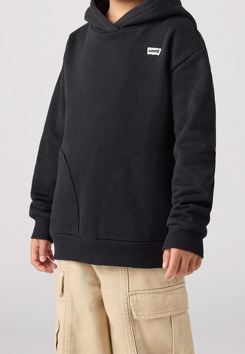 Enfant portant un sweat à capuche Levi's noir avec poches avant et un pantalon cargo beige, montré du cou au milieu de la cuisse sur un fond uni.