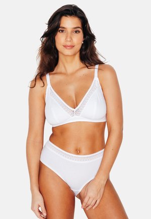 Donna con lunghi capelli scuri che indossa un reggiseno bianco con rifiniture in pizzo e biancheria intima a vita alta, in piedi davanti a uno sfondo bianco uniforme.