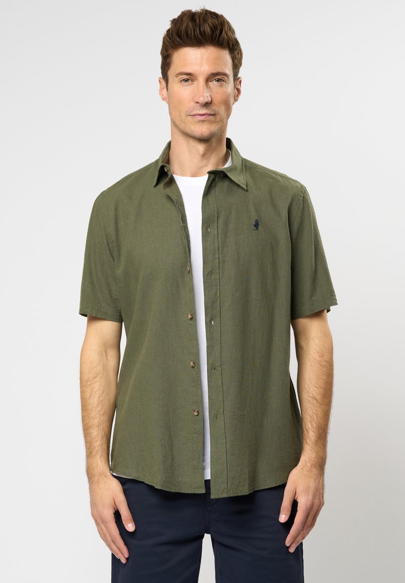 Camicetta a maniche corte di colore verde oliva con tessuto testurizzato, apertura con bottoni e un piccolo logo ricamato sul petto. Indossata sopra una t-shirt bianca.