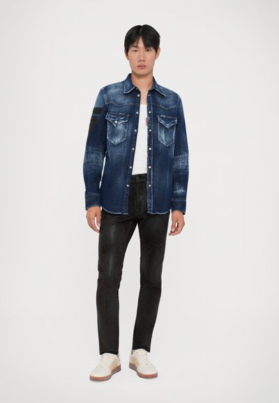 Camicia in denim di colore blu scuro con leggera abrasione, tasche sul petto doppie e toppe sui gomiti, abbinata a pantaloni neri e sneakers bianche.
