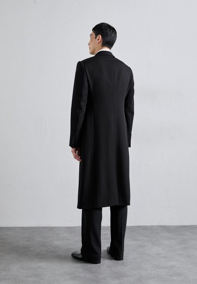 Melnā gara mētela, kas izgatavota no teksturēta auduma, ar pielāgotu piegriezumu, izgrieztiem atlokiem un pogājamiem piedurknēm, apvienota ar atbilstošām biksēm un kurpēm.