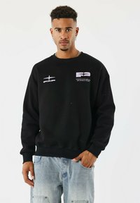 Homme avec des boucles d'oreilles et des cheveux bouclés courts portant un sweat noir avec des logos et un jean baggy bleu clair, se tenant avec une main dans la poche.