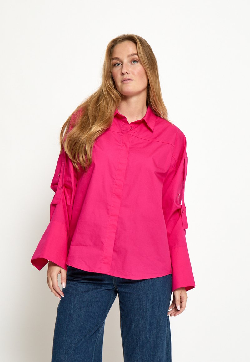 Mujer con cabello largo y rubio, vistiendo una camiseta de manga larga rosa brillante con puños anchos y vaqueros azul oscuro, de pie contra un fondo blanco.