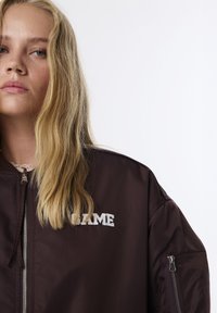 Cazadora bomber marrón con el logo "GAME" bordado en blanco, cierre frontal con cremallera y cremalleras en contraste en las mangas. Textura de tela suave y brillante.