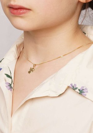 Goldene Halskette mit einem kleinen Anhänger in Form eines Schmetterlings, getragen über einem cremefarbenen Hemd mit Kragen und lila floralem Stickmuster.