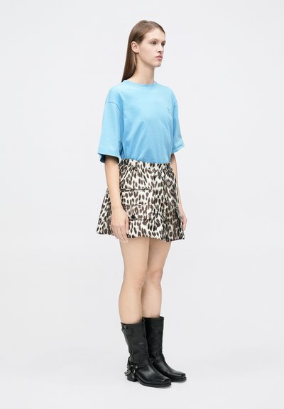 Jeune femme debout, portant un t-shirt oversized bleu clair, une mini-jupe à motif léopard avec des poches cargo, et des bottes en cuir noires mi-mollet.