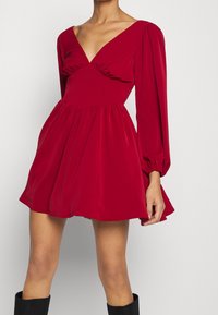 Rotes Mini-Kleid aus glattem Stoff, mit tiefem V-Ausschnitt, Puffärmeln und einer gerafften Taille mit einem ausgestellten Rock. Schwarze kniehohe Stiefel.