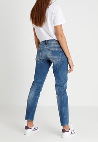 Jeans de mezclilla azul con un corte ajustado, que cuentan con bolsillos traseros y un ligero desgaste. Combinados con una camiseta blanca y zapatillas con detalles morados.