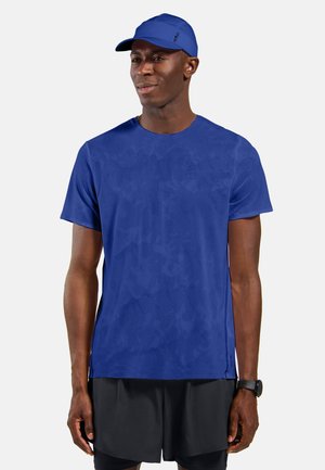 Man draagt een blauwe sportpet en bijpassend blauw gestructureerd t-shirt met zwarte shorts tegen een effen achtergrond.